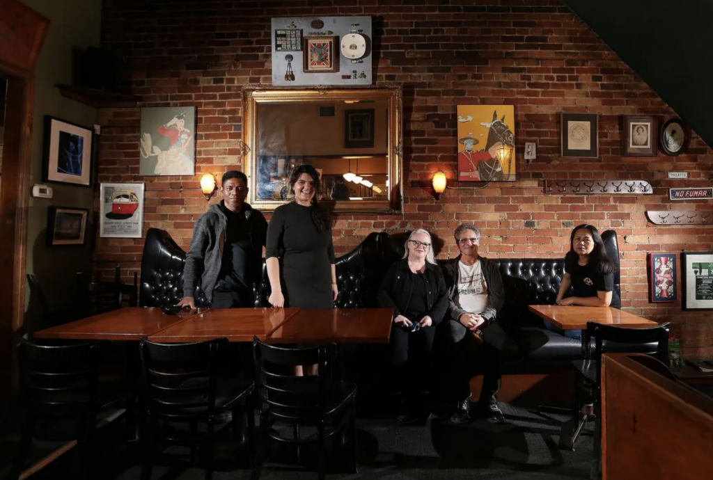 Owners Andre and Kelly with long time staff and now business partners, Sarah and Ranjan, and Chef Noy in the dining room, against a brick wall with leather banquet seating